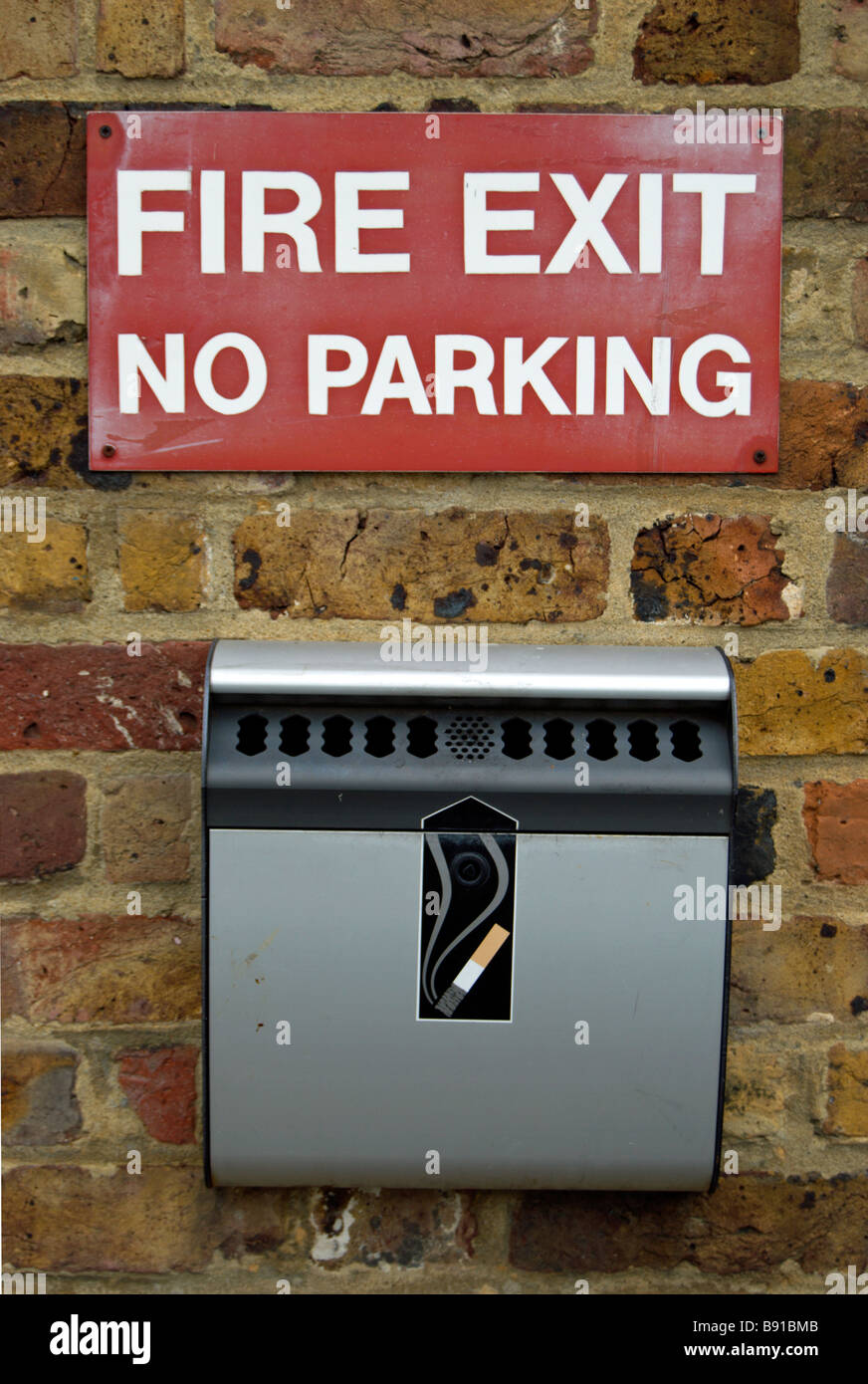 fire exit and no parking sign above a cigarette bin with illustration of a lit cigarette, set against a brick wall Stock Photo