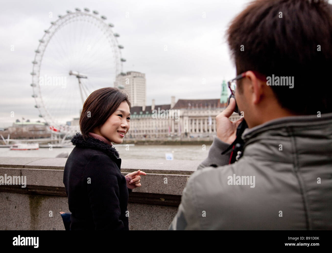 Asian oriental tourist couple in 20s / 30s visiting London, he taking photo of her in front of London Eye Stock Photo