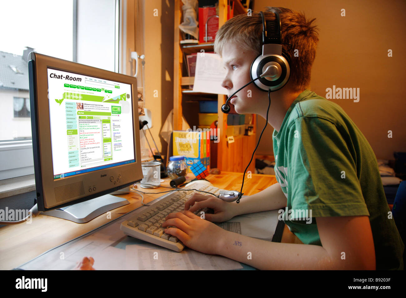 A boy, 13 years old, is chatting in his child room and talks to a chat member by voice over ip. He wears a headset. Stock Photo