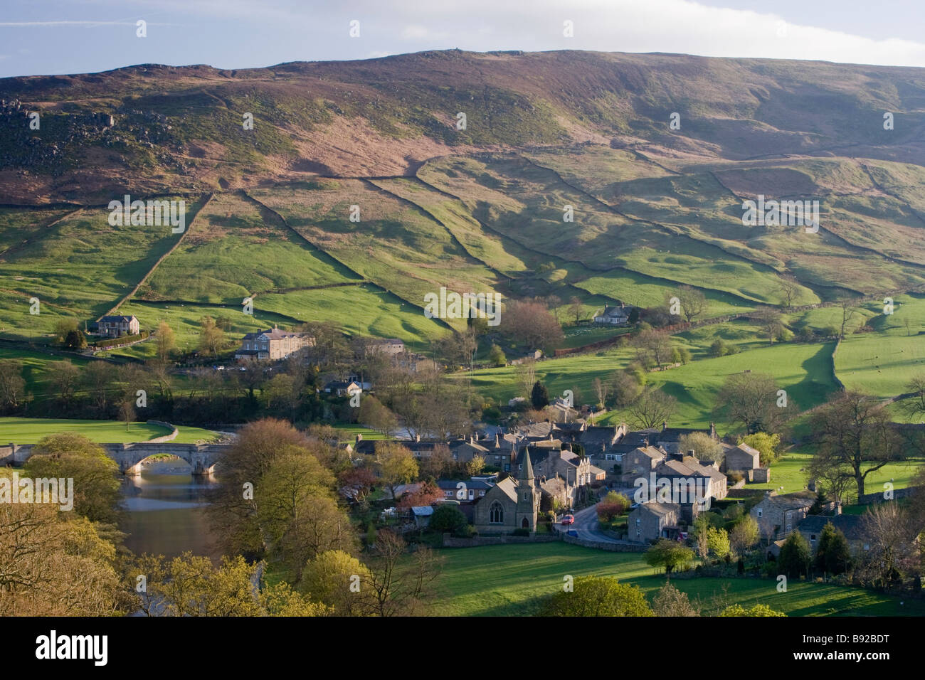 High view over beautiful, sunny Yorkshire Dales village of Burnsall in valley by River Wharfe & with steep, green, hillside beyond - England, GB, UK. Stock Photo