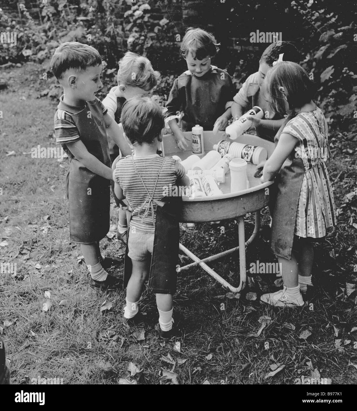 1950s, early learning, a group of young children playing with old washing-up liquid plastic bottles in a metal bowl outside in a garden, England, UK. Stock Photo