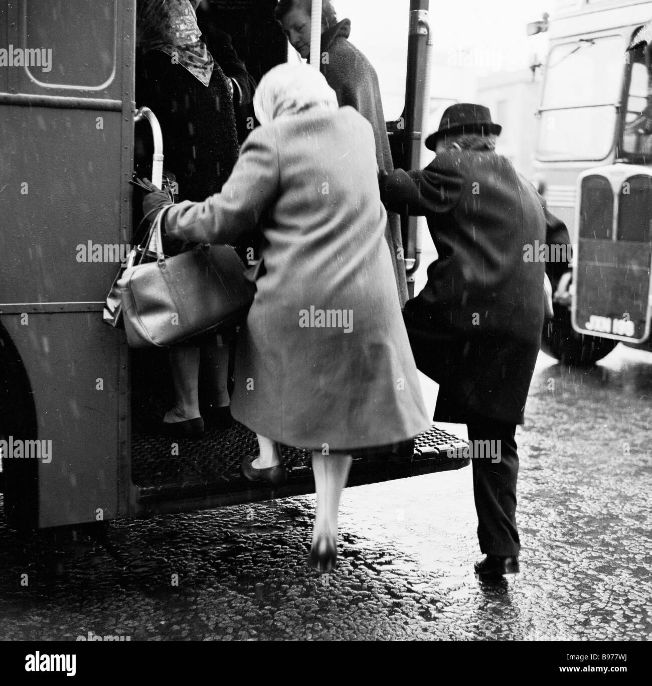 1950s, winter time, an elderly man and woman grap the hand-rails and step up onto the open rear platform of a double-decker bus, London, England, UK. Stock Photo