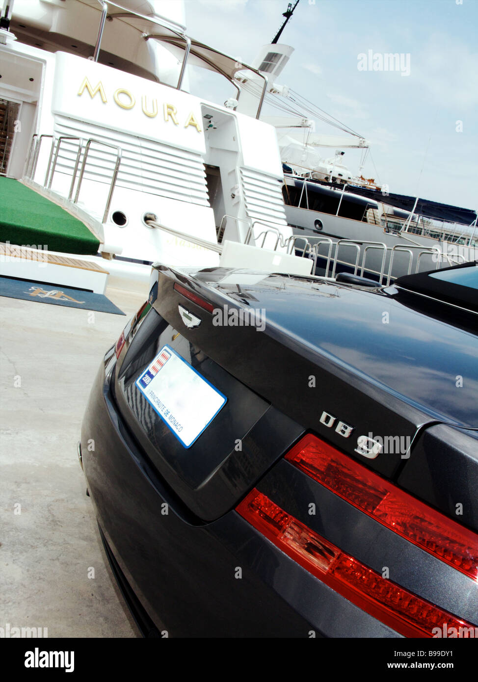 A blue Aston Martin DB9 parked alongside the Lady Moura luxury super yacht moored in the port of Monaco Monte Carlo. Stock Photo