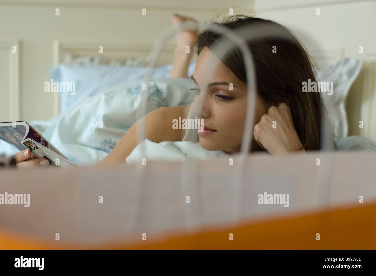 Young woman lying in bed reading magazine, shopping bag in foreground Stock Photo