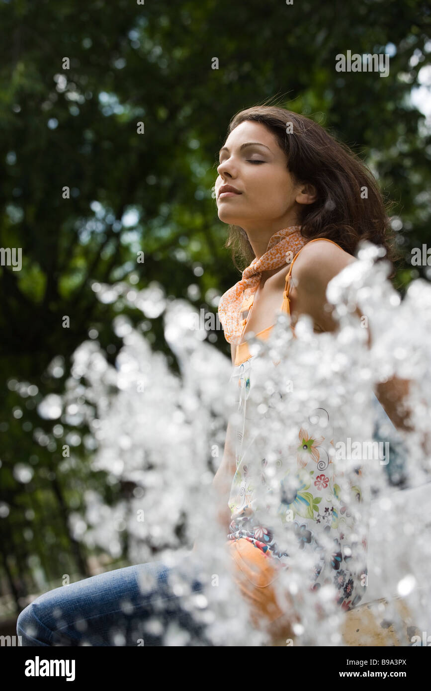 Young woman sitting by outdoor fountain with eyes closed, low angle view Stock Photo