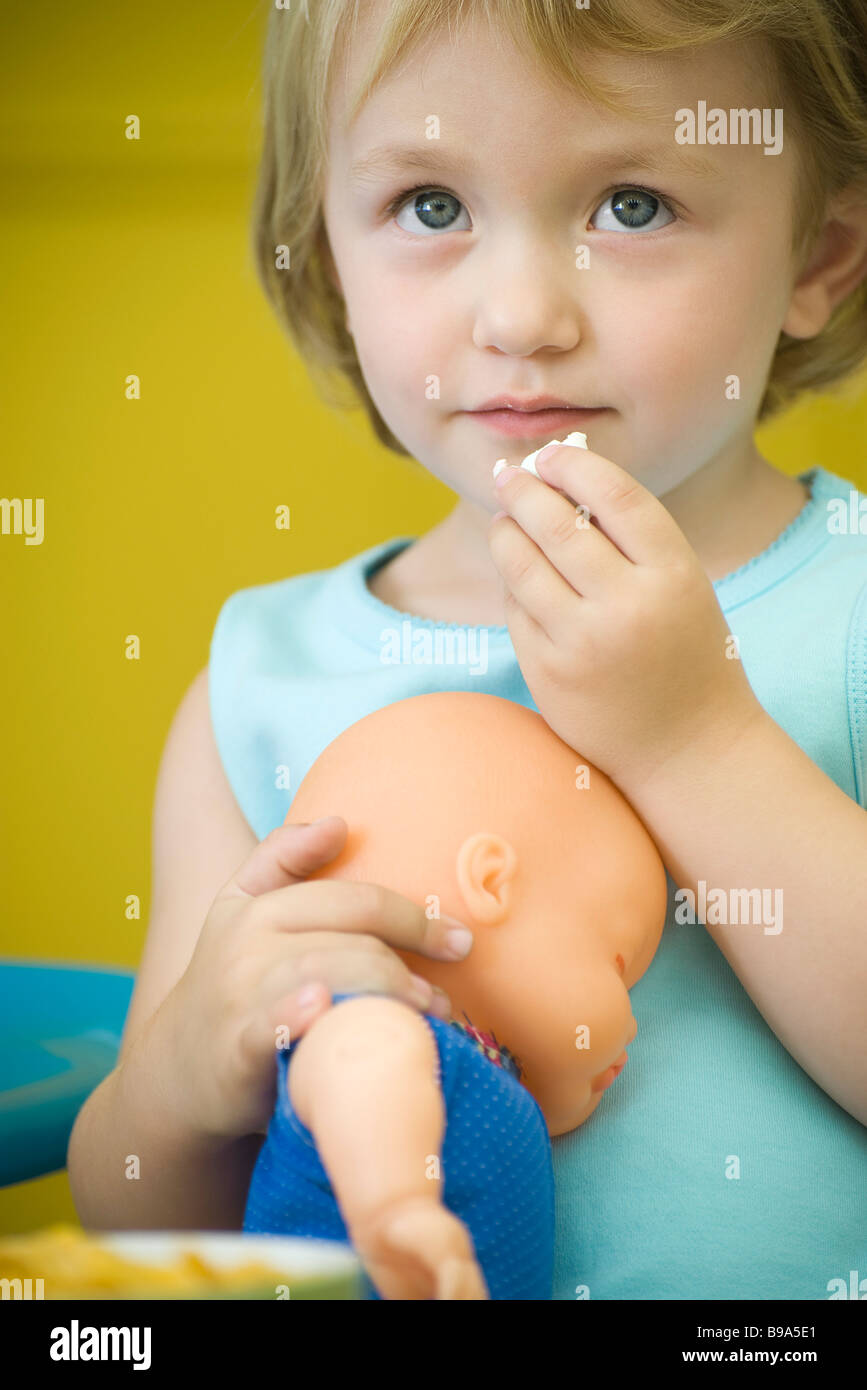 Little girl holding baby doll, eating snack Stock Photo - Alamy