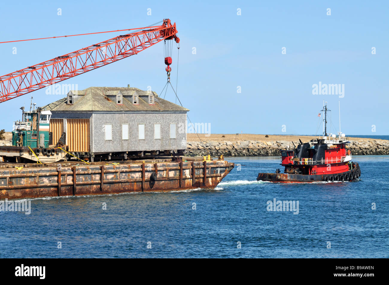 Barge moving a wooden house with small pushy tug and crane loaded onboard being pulled by tugboat through the Cape Cod Canal in Massachusetts USA Stock Photo
