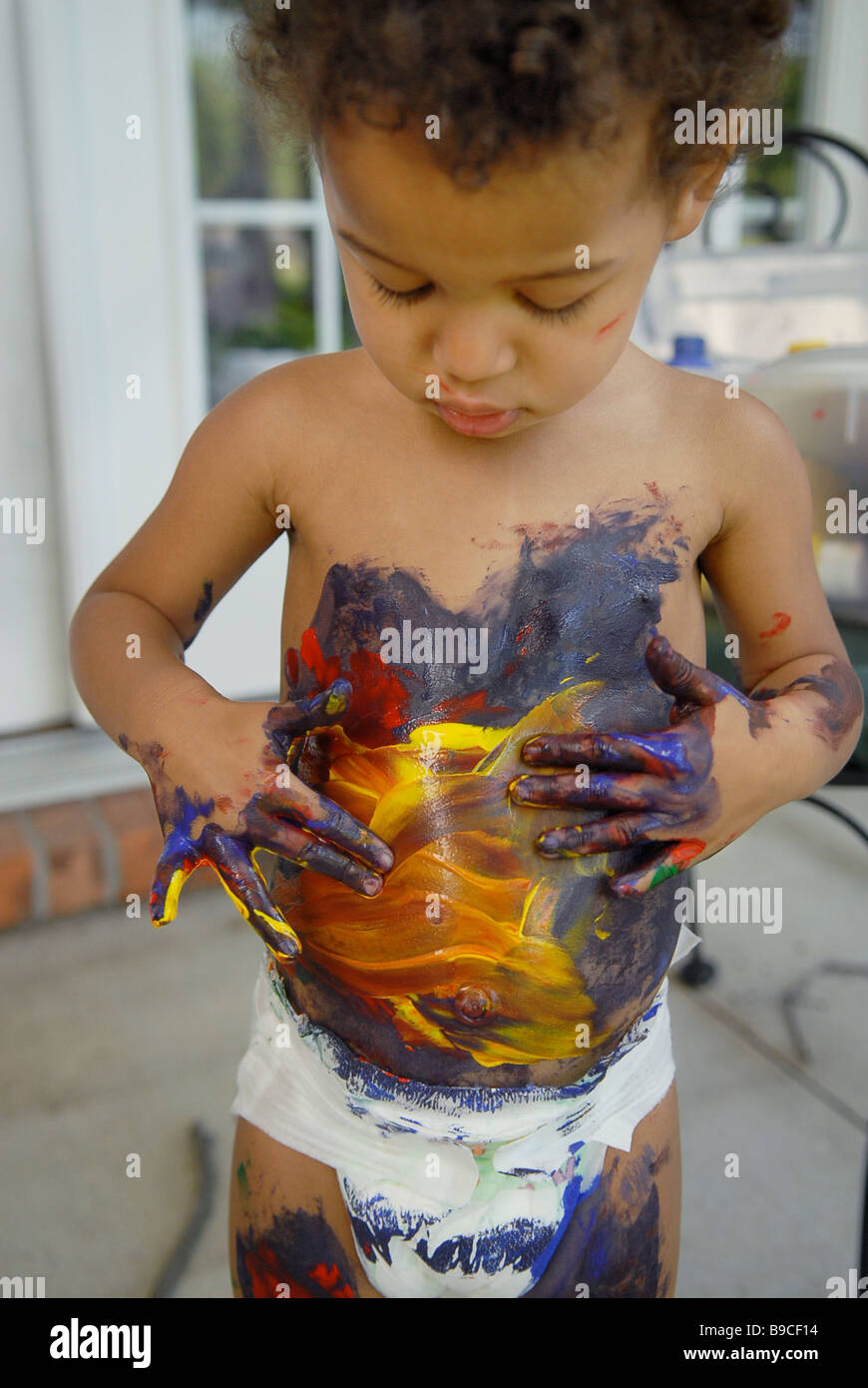 An African American biracial child paints her body with red green blue and yellow primary colors as she plays outside Stock Photo