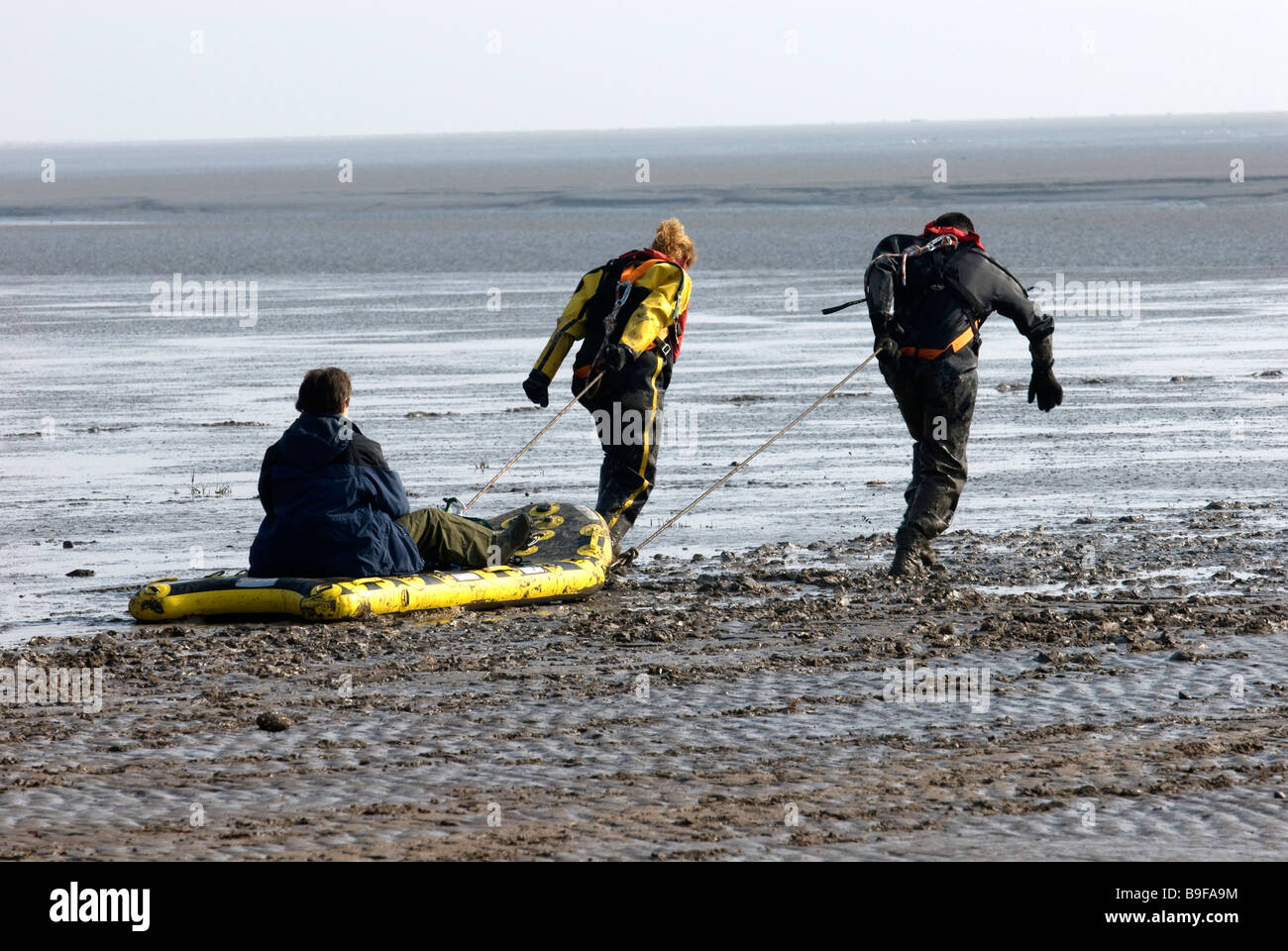 Two Coastguard Rescue Personnel transport a woman on an inflatable skid across mud flats on beach Stock Photo