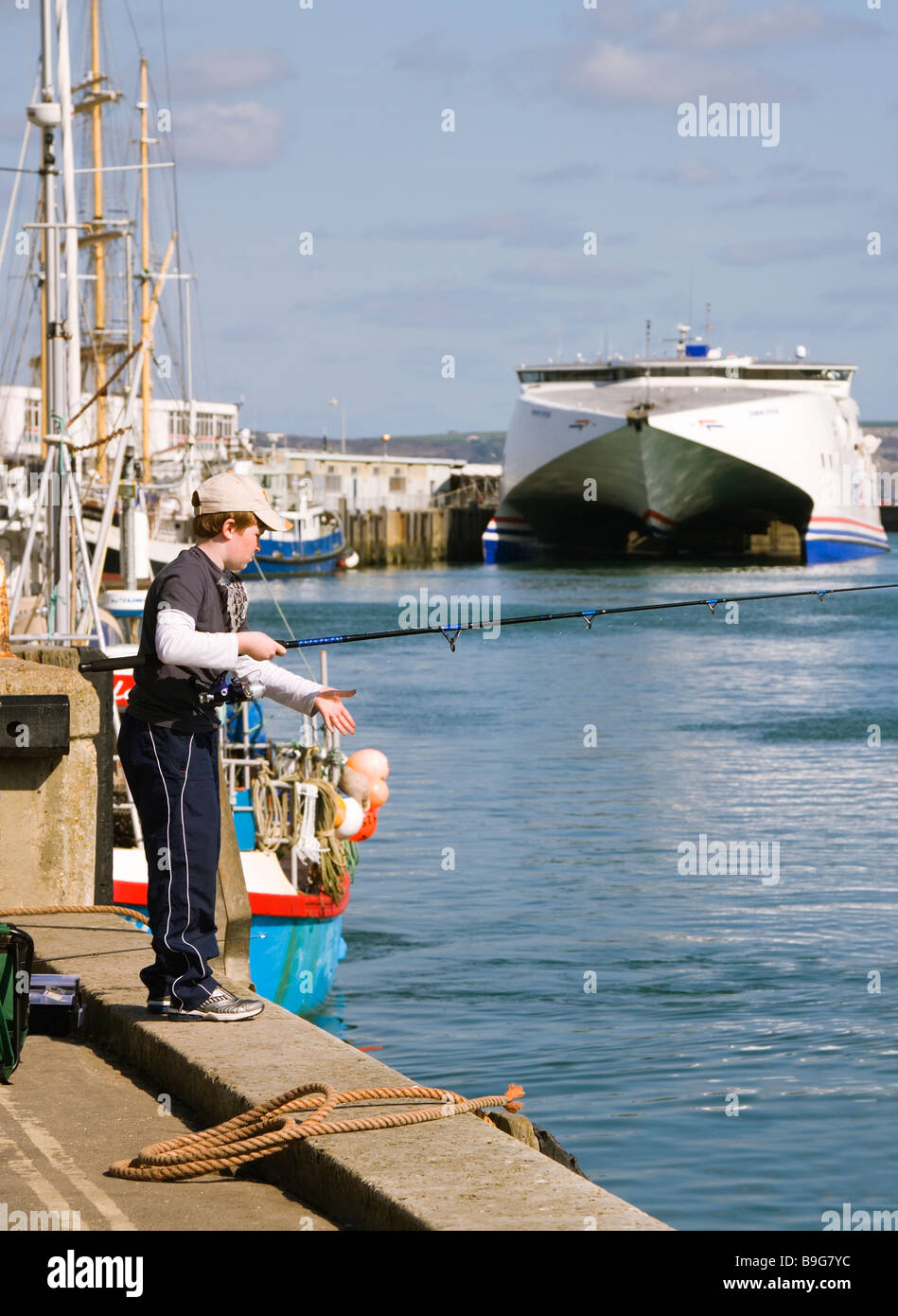 A young boy fishing off Weymouth harbour wall. Dorset. UK.  Cross channel ferry, the Condor Ferries catamaran at berth. Stock Photo