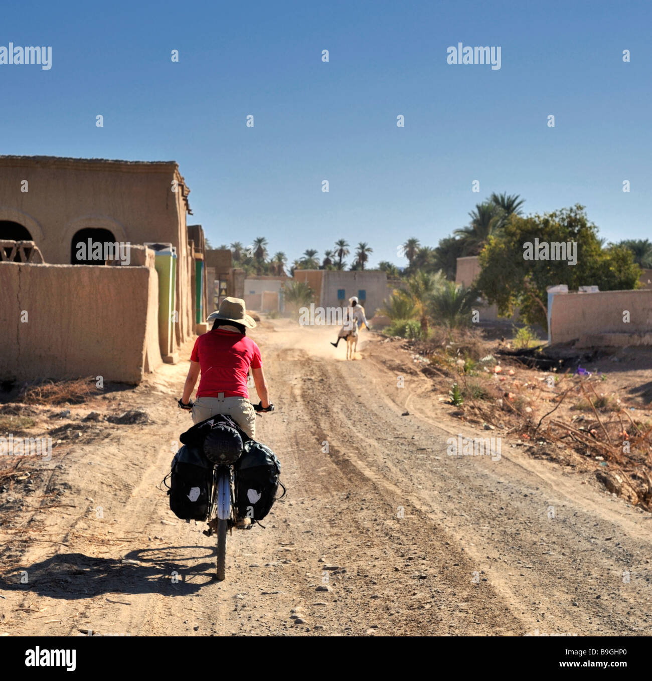 western female touring cyclist going away from camera into Nubian village of northern Sudan with man on donkey in distance Stock Photo