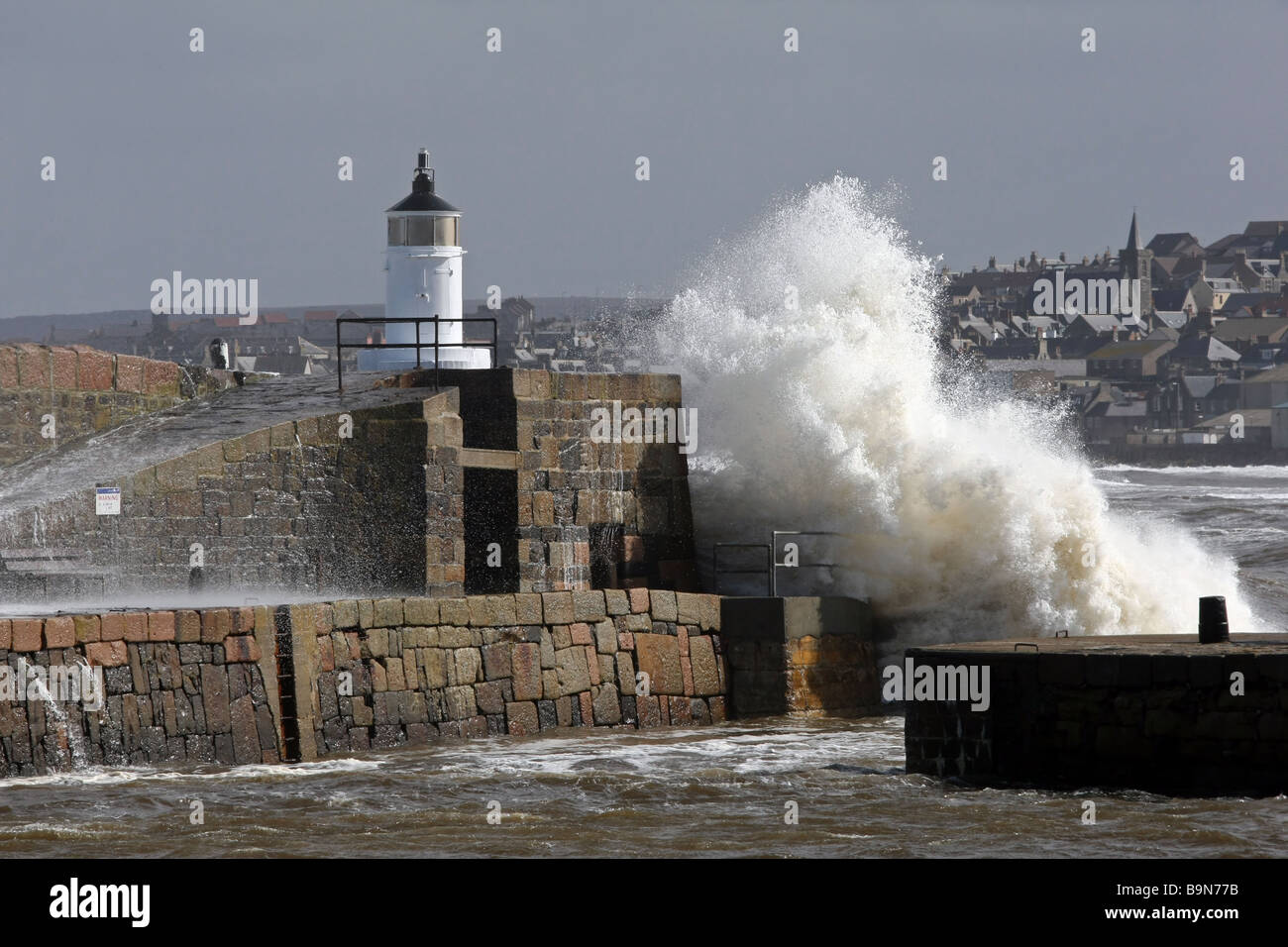 Waves break over the wall and lighthouse at the entrance of Banff Harbour, Scotland, UK. Town of Macduff is in the background Stock Photo