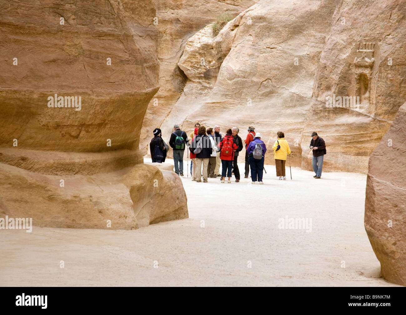 Tourists walking through the siq, past ancient wall carvings, towards the entrance to Petra, Jordan Stock Photo