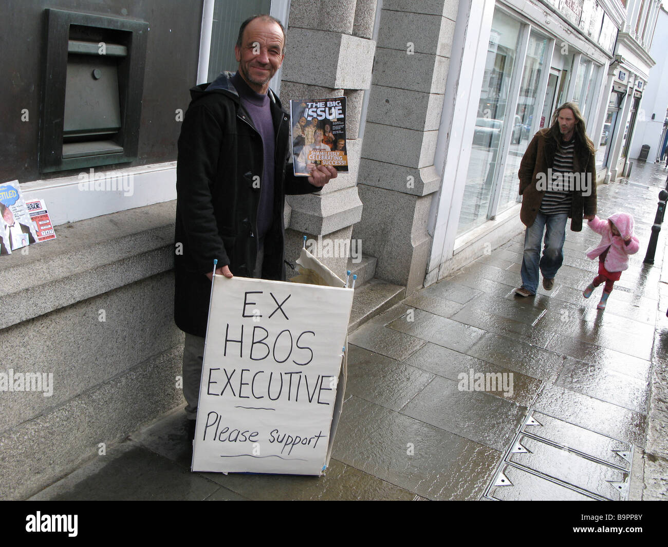 A Big Issue Seller with a spoof billboard saying 'Ex HBOS executive Please Support' next to a bank ATM hole in the wall. Stock Photo