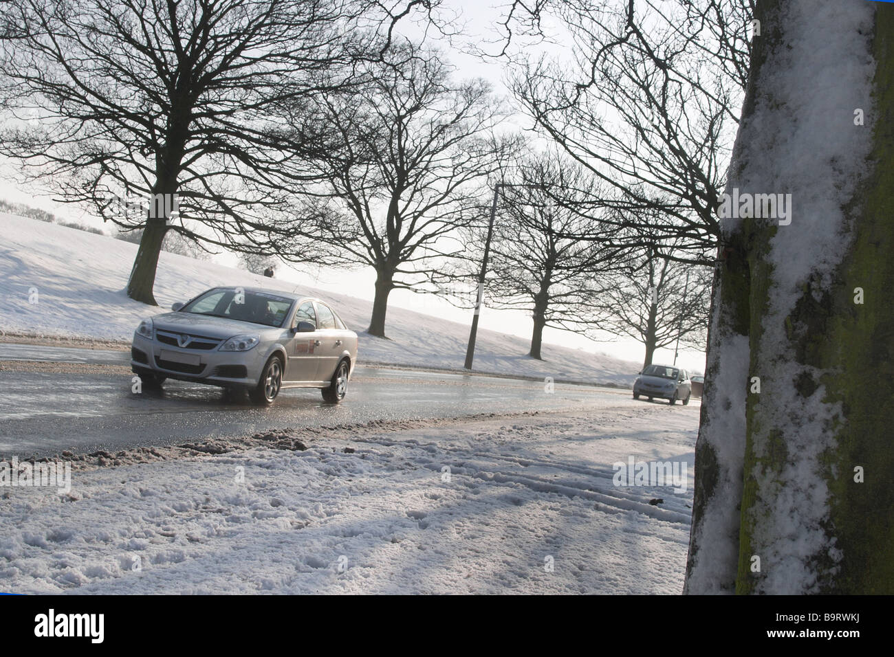 Snow scene with trees and road running through and cars travelling causing water spray. shot in strong sunlight causing shadows Stock Photo