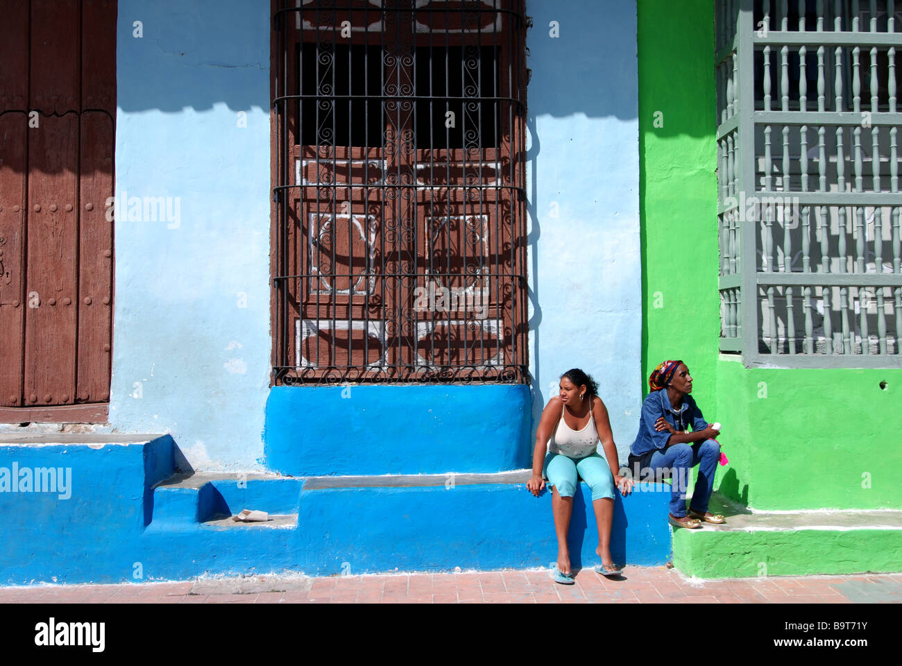 Two women sitting on stair in front of houses qith gated windows Camaguey Cuba Stock Photo