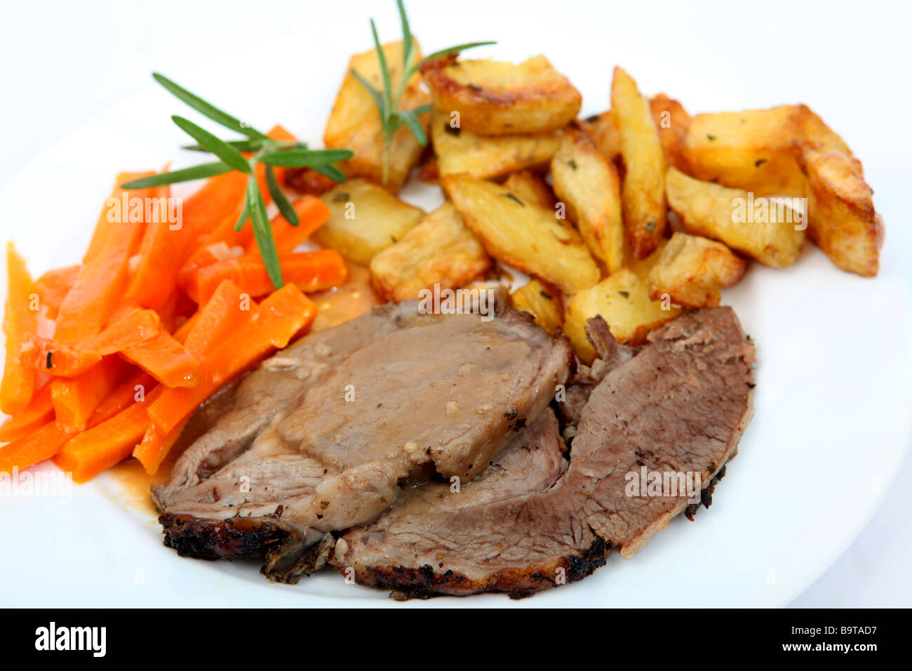 Slices of herbed roast lamb with gravy served with steamed garlic and carrots and diced roast potatoes and sprigs of rosemary Stock Photo