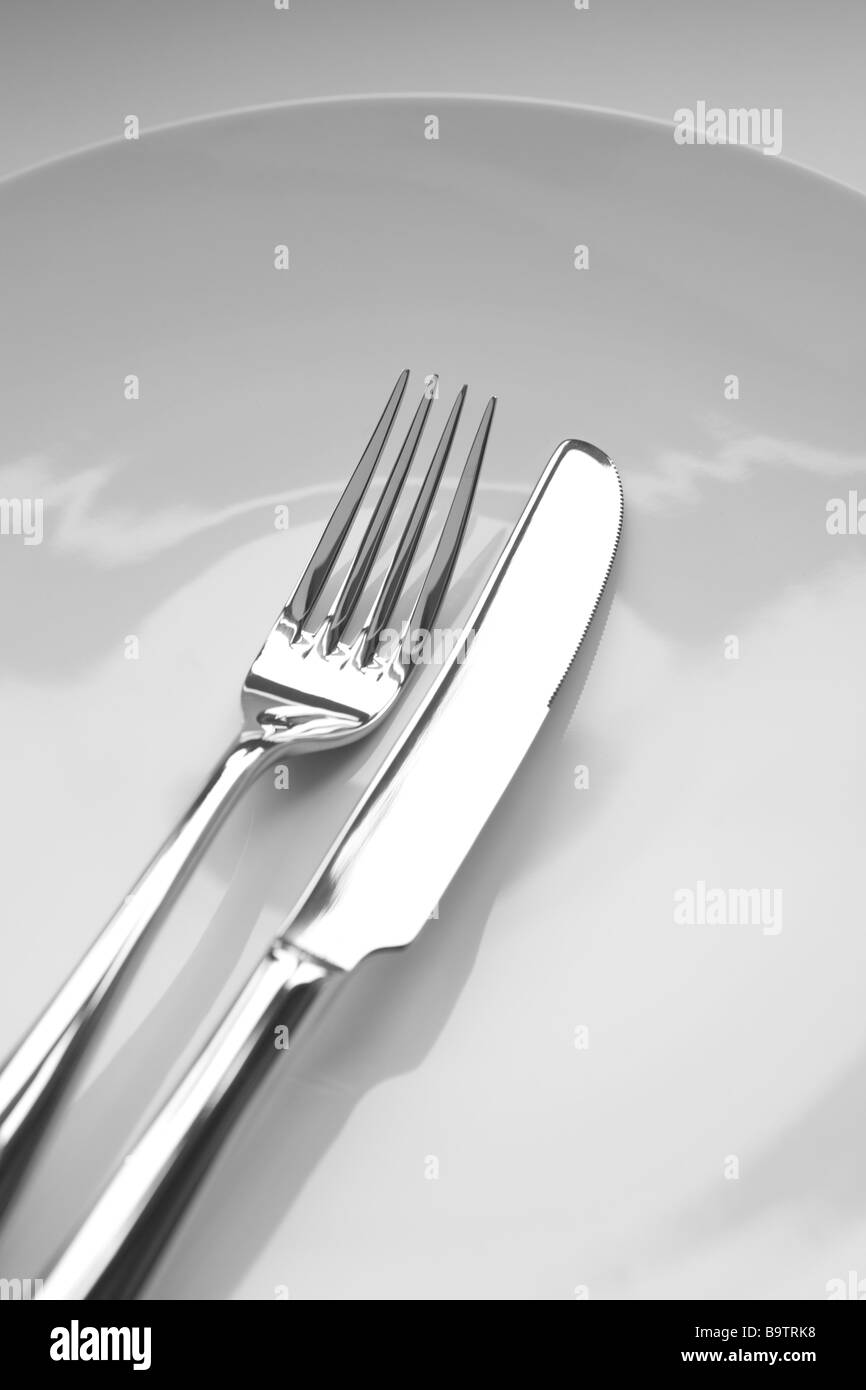 stainless steel knife and fork on a white plate and background. Narrow depth of field Stock Photo