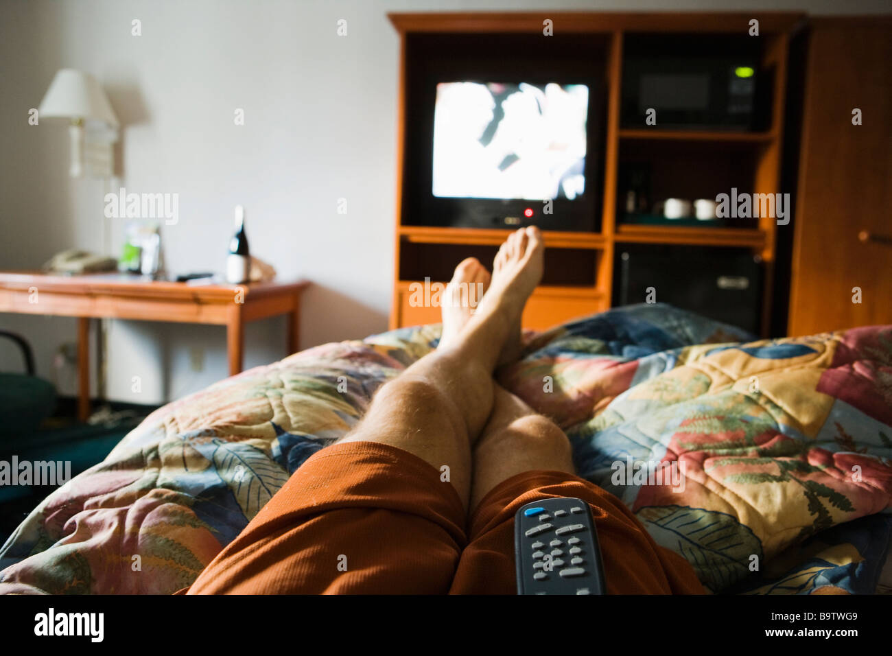 A first peson view of a mans legs relaxing on a bed in a hotel room watching the TV with a remote control on his lap Stock Photo