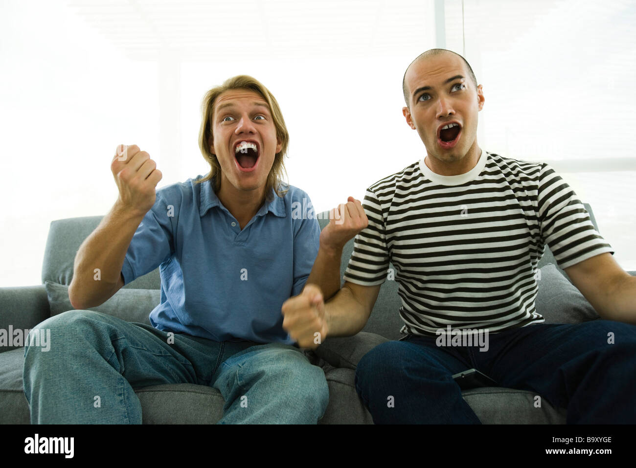 Two men sitting on sofa watching TV, cheering with clenched fists Stock Photo