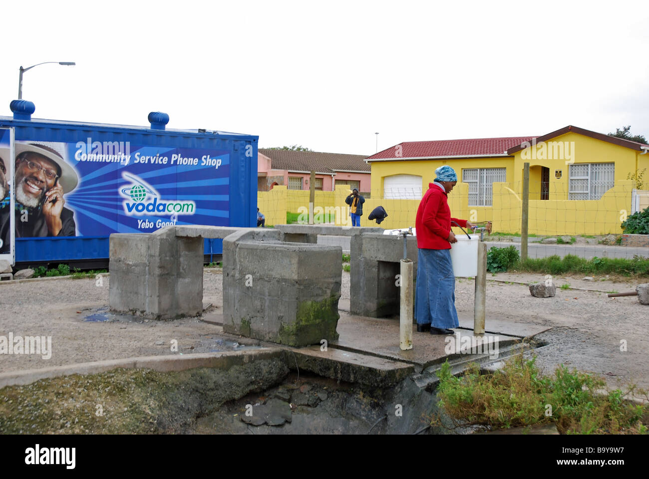 A young woman in an area of informal housing fills a bucket from a communal standpipe, Langa Township, Cape Town, South Africa Stock Photo