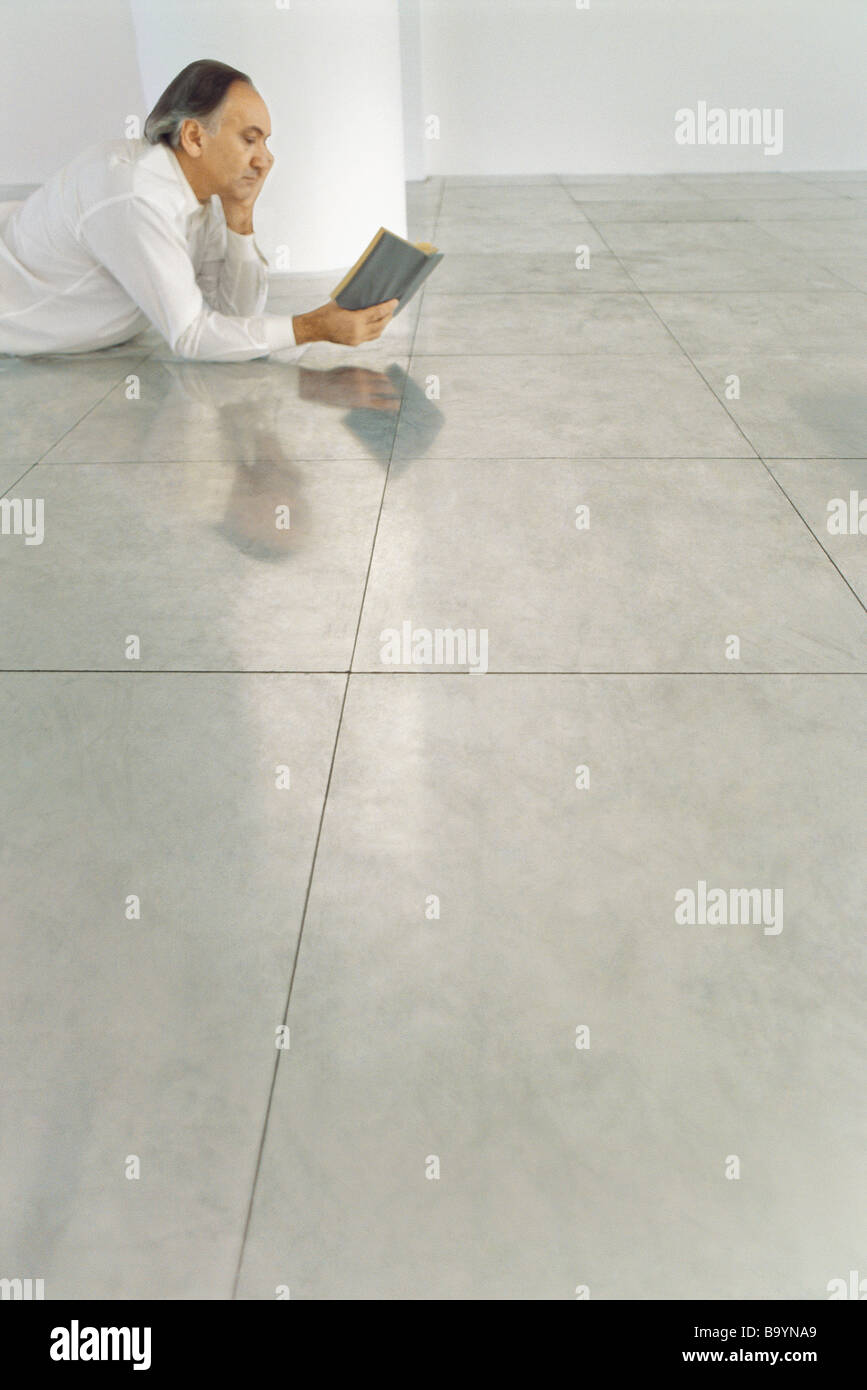 Senior man lying on stomach on tiled floor reading book Stock Photo