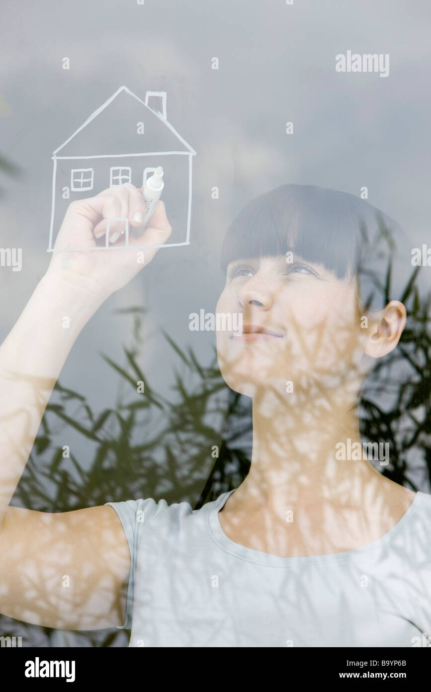 Young woman drawing house on windowpane, daydreaming Stock Photo
