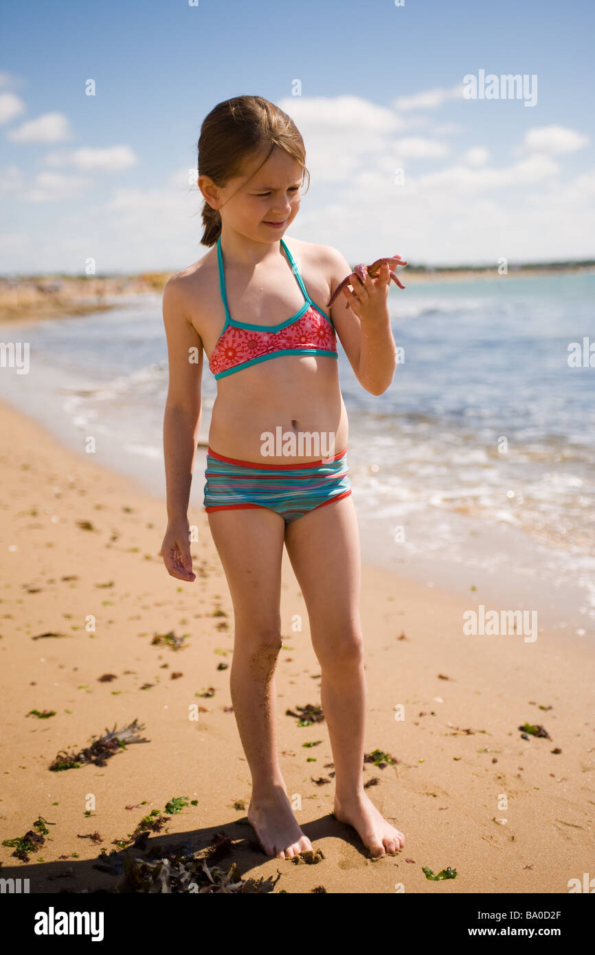 A girl holds a starfish in her hand to examine it on a beach with waves washing in behind her on a sunny day at the beach. Stock Photo