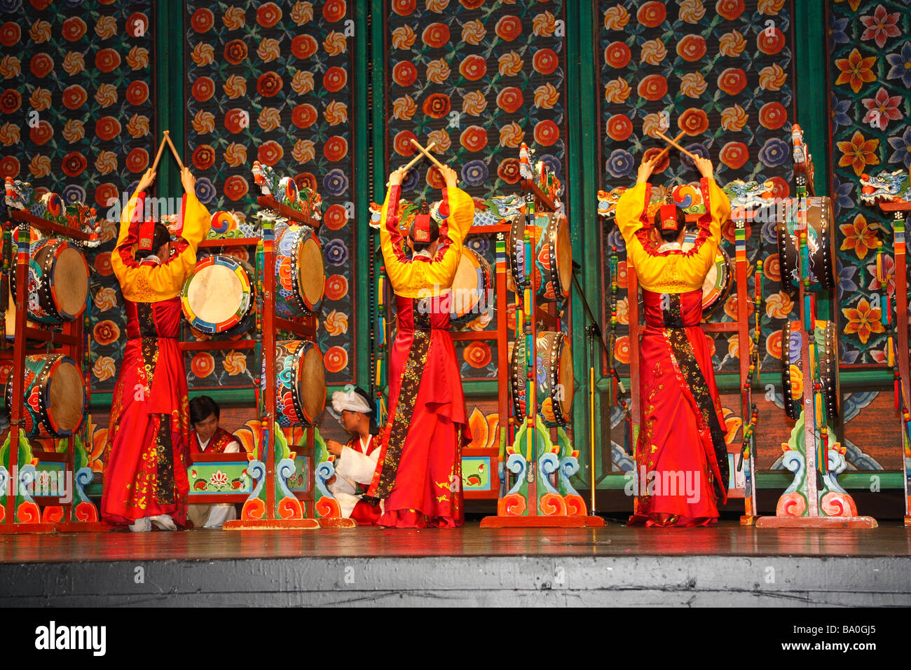 Three Korean women in bright yellow and red dresses performing on stage playing traditional folk drums 'oh-go-mu' Stock Photo