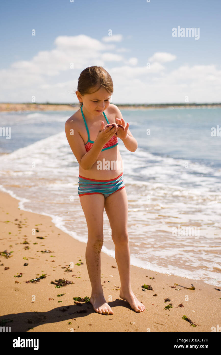 A girl holds a starfish in her hand to examine it on a beach with waves washing in behind her on a sunny day at the beach Stock Photo