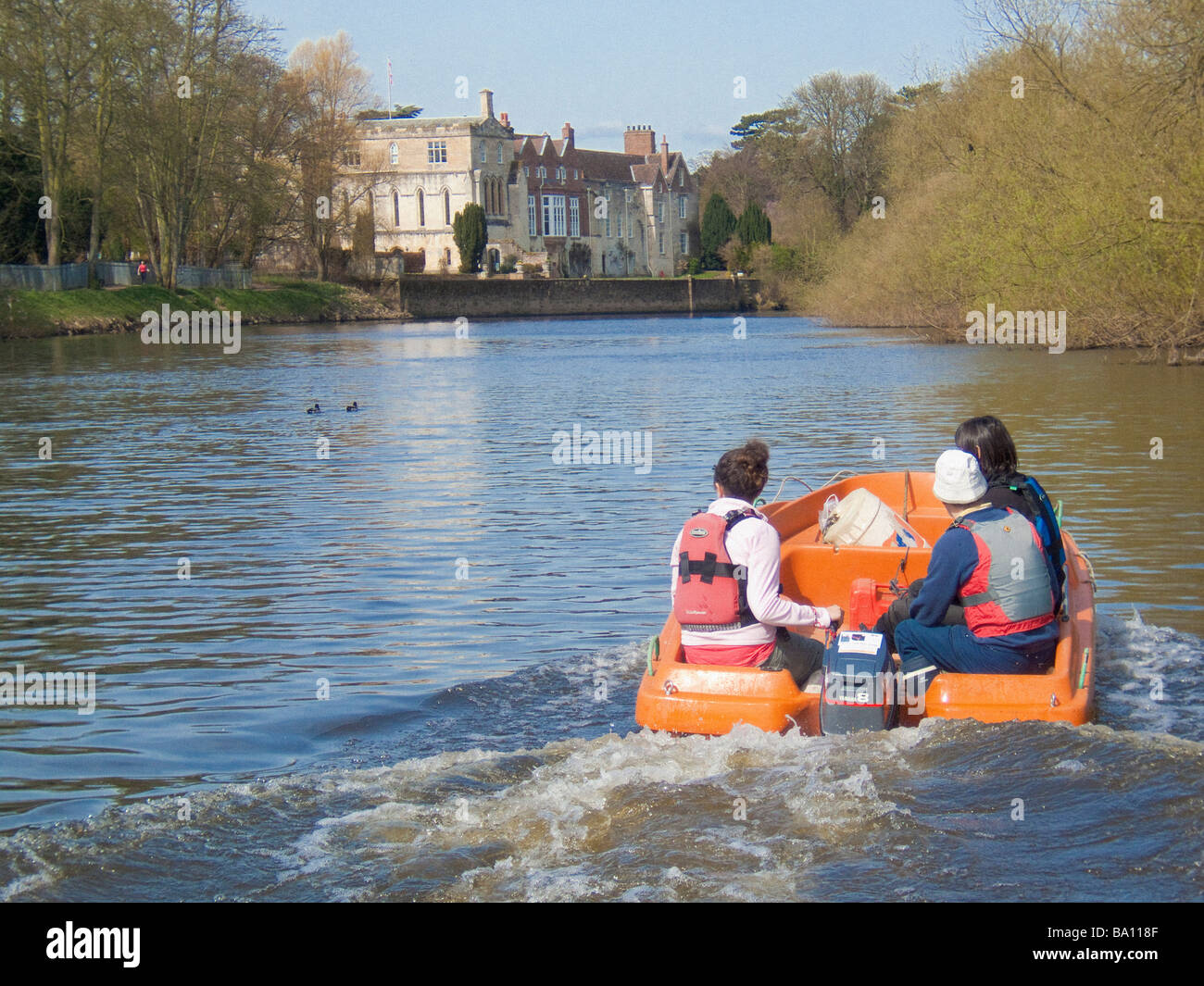 Orange plastic moulded boat with 3 people on board, on the river Ouse in York with Bishopthorpe Palace in the distance. Stock Photo