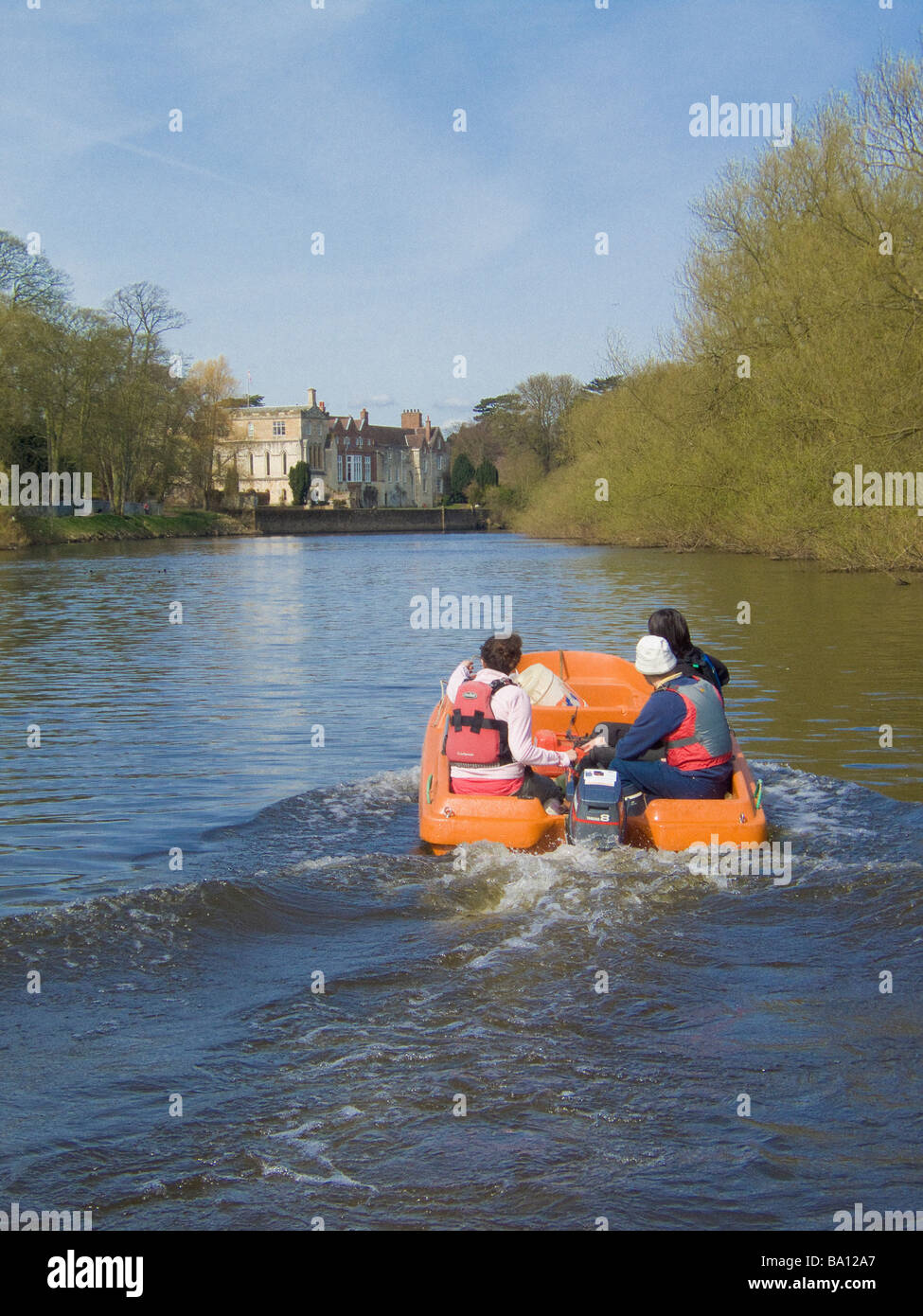 Orange plastic moulded boat with 3 people on board, on the river Ouse in York with Bishopthorpe Palace in the distance. Stock Photo