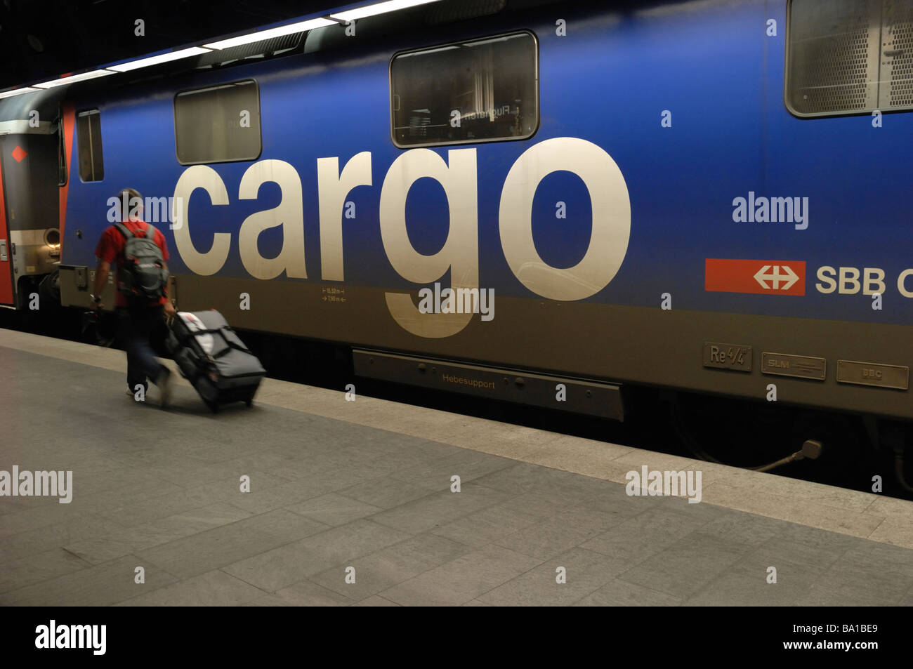 A passenger pulling a wheely bag on the train platform below Zurich Airport in Switzerland as a freight train goes past Stock Photo