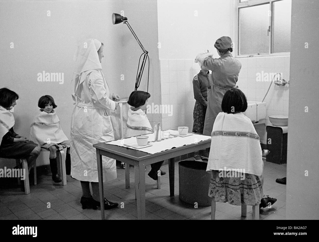 1950s, children with tea towels on their shoulders wait in turn to have their hair inspected for lice or nits by the local district nurse, England, UK Stock Photo