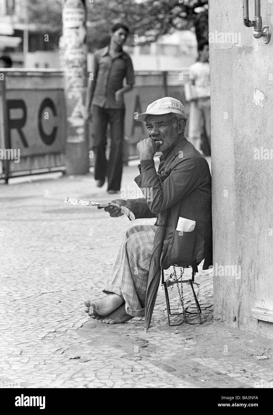 Seventies, black and white photo, people, poorness, gambling, older man sits at a corner of a house selling lottery tickets, barfooted, tattered clothing, aged 60 to 70 years, Brazil, Minas Gerais, Belo Horizonte Stock Photo