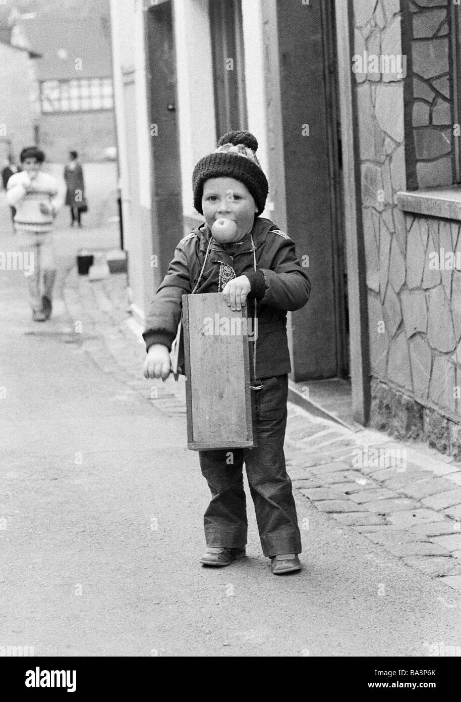 Seventies, black and white photo, people, children, little boy with a ratch or rattle, apple in the mouth, aged 4 to 7 years, Easter Customs in the Eifel region, in the Holy Week in rural areas local children walk around with ratches or rattles, symbolically this rasping or rattling shall replace the peal of bells because in the Passion Week all bells had been travelled to Rome, D-Monreal, Elz, Elzbach, Elz Valley, Eifel, Verbandsgemeinde Vordereifel, Rhineland-Palatinate Stock Photo