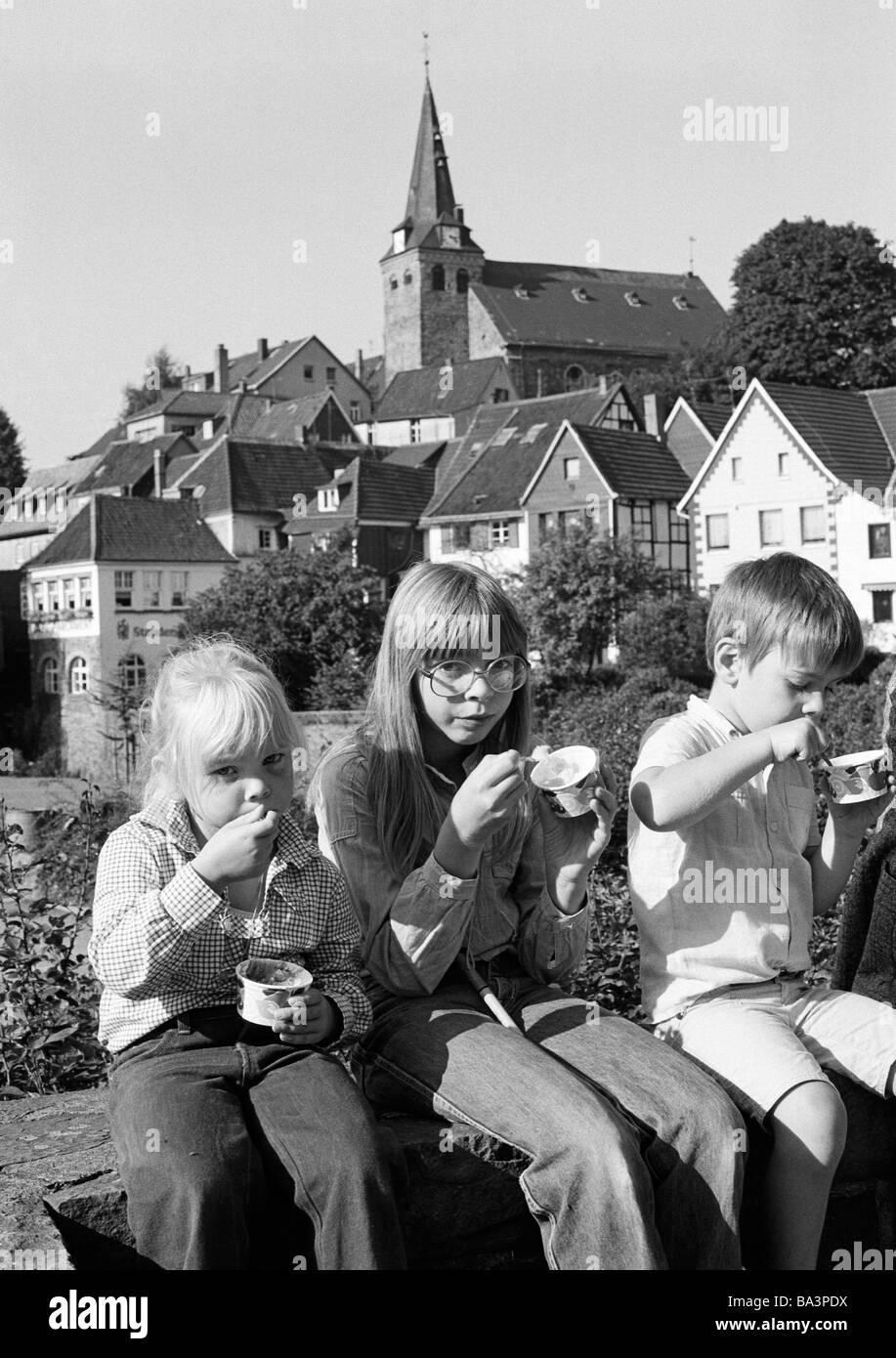 Seventies, black and white photo, people, children, two girls an a boy sit on a wall nibbling ice cream, aged 4 to 12 years, in the background the old town of Kettwig, evangelic market church, residential buildings, D-Essen, D-Essen-Kettwig, Ruhr area, North Rhine-Westphalia Stock Photo