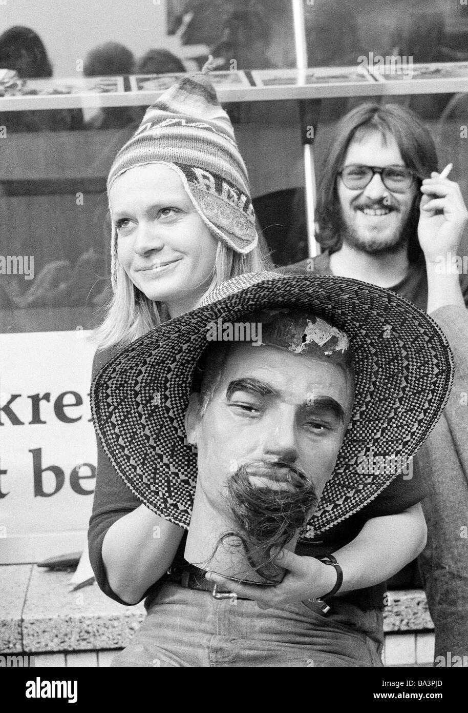 Seventies, black and white photo, humour, people, flea market, young woman presents a procuct, mans head with strawhat, doll, puppet head, young man in the background laughs, aged 20 to 25 years Stock Photo