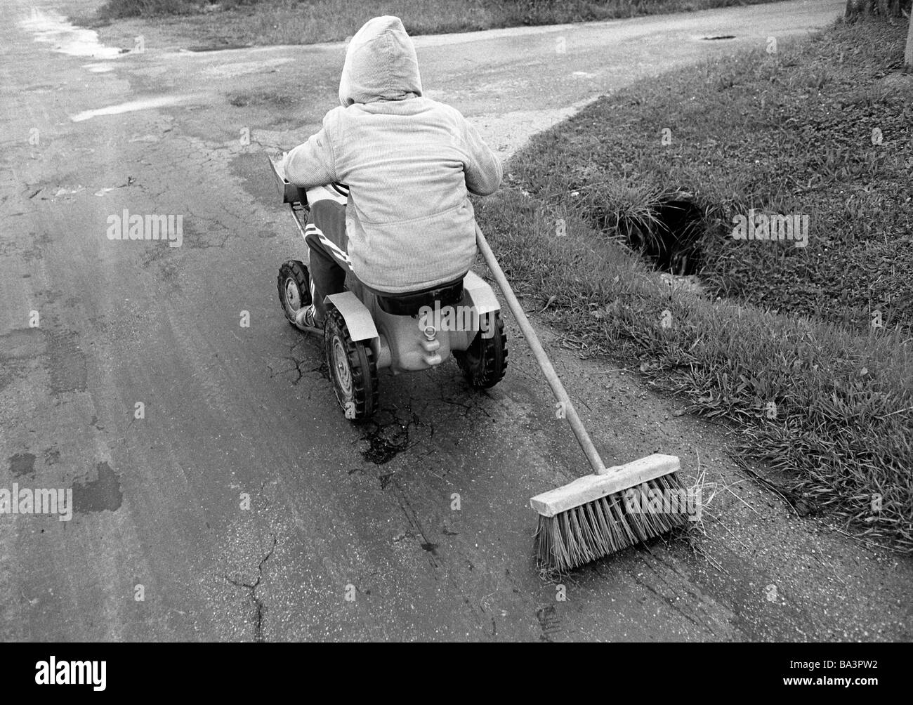 Eighties, black and white photo, people, children, little boy drives in a toy car along the street pulling a besom, street sweeper, aged 4 to 6 years Stock Photo