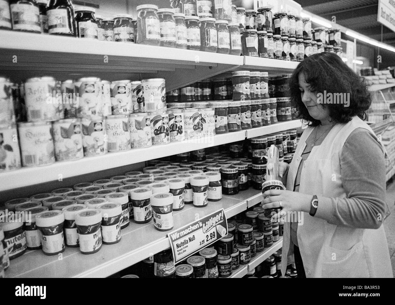 Eighties, black and white photo, people, working life, retailing, shop, viands, salesgirl, young woman in a work coat stands near a grocery shelf and prices the goods by a labeling machine, aged 23 to 30 years Stock Photo