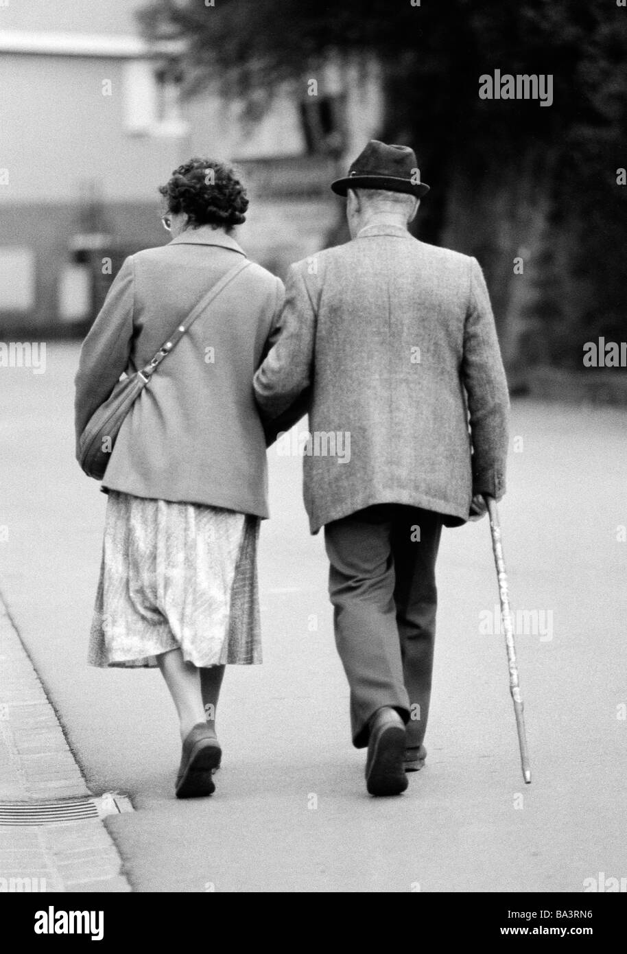Eighties, black and white photo, people, elder people, older couple walking arm in arm, jacket, trousers, skirt, hat, aged 65 to 75 years Stock Photo