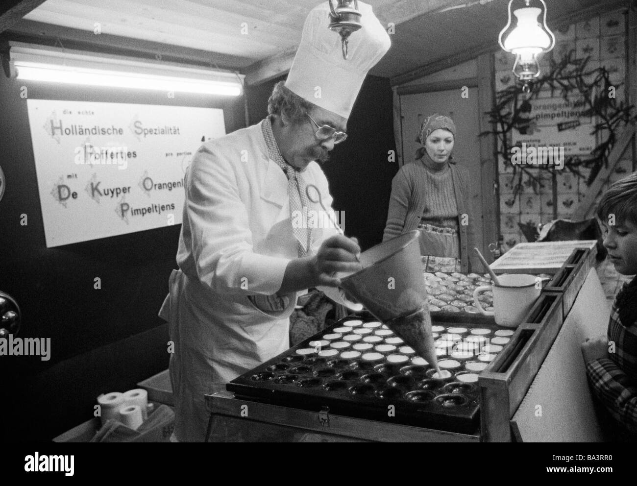 Seventies, black and white photo, people, kermess, funfair, gourmet stall, cook with chefs hat prepares to bake sweet cookies and cakes, Poffertjes are a speciality from the Netherlands, aged 40 to 50 years, in the background a femal coworker wearing a headscarf, aged 30 to 40 years Stock Photo