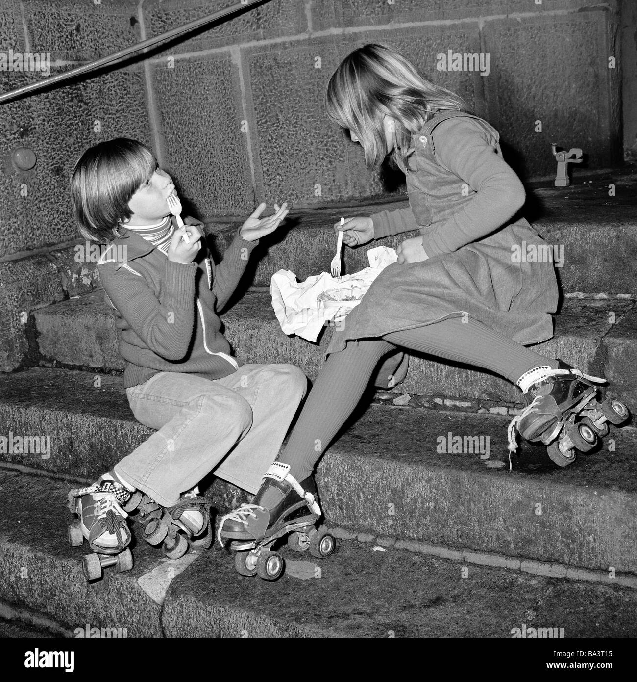 Seventies, black and white photo, people, children, two little girls with strapped rollerblades sit in a hall on a stairs and eat fastfood, aged 8 to 12 years Stock Photo