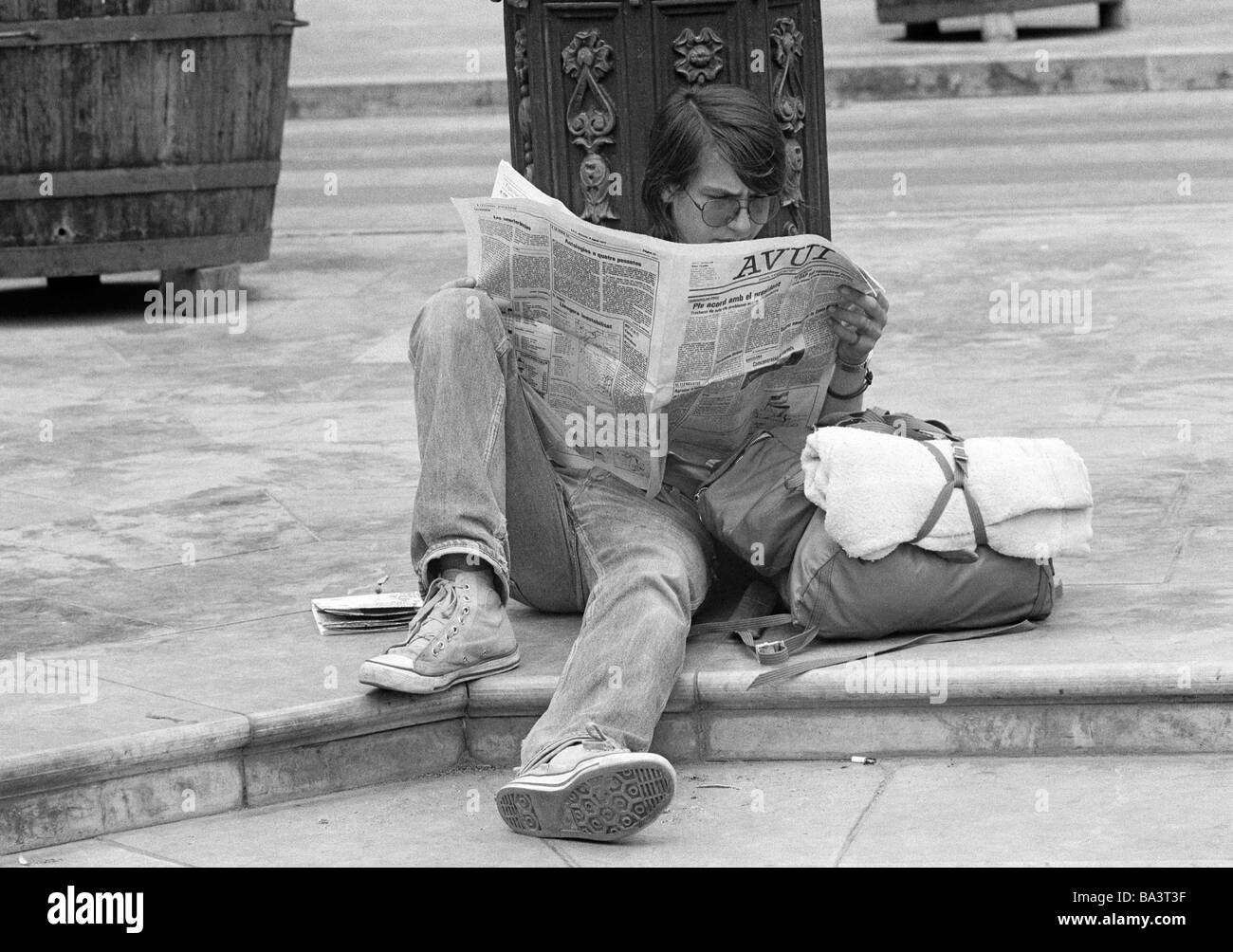 Seventies, black and white photo, people, teenager sits at the roadside reading a newspaper, luggage, bag, jeans trousers, plimsolls, aged 18 to 22 years, Spain, Valencia Stock Photo