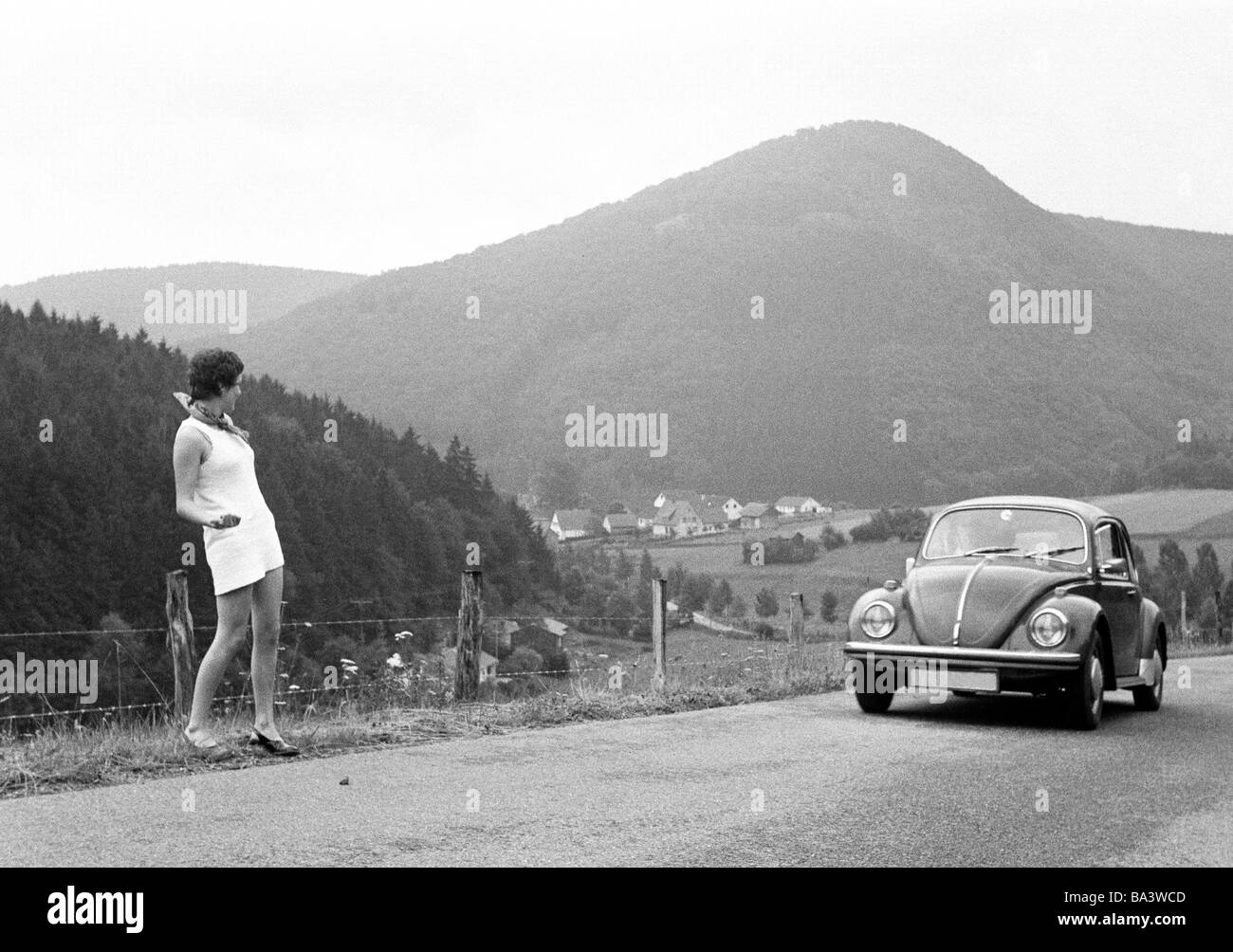 Seventies, black and white photo, holidays, travel, hitchhiker, young girl in a mini dress stands on the roadside of a country road in a mountainous landscape requesting to be given a lift by an automobile type VW beetle riding past, aged 17 to 23 years, Monika, D-Brilon, Sauerland, North Rhine-Westphalia Stock Photo