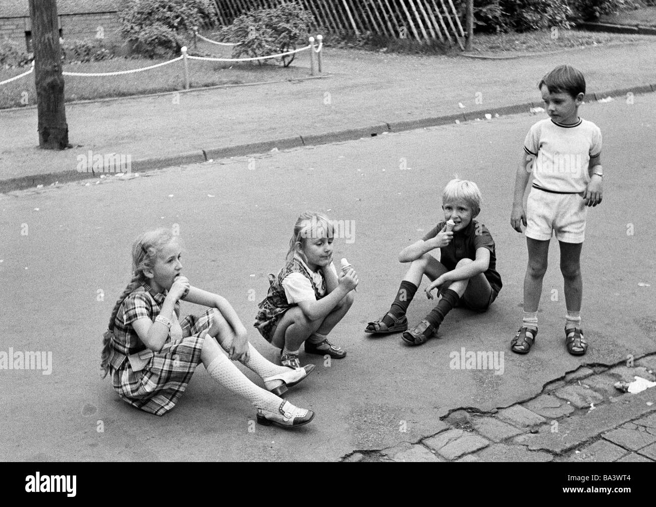 Seventies, black and white photo, people, children, two girls and a boy sitting on a street and lick ice cream, another boy stands beside them and looks envious, aged 5 to 9 years Stock Photo