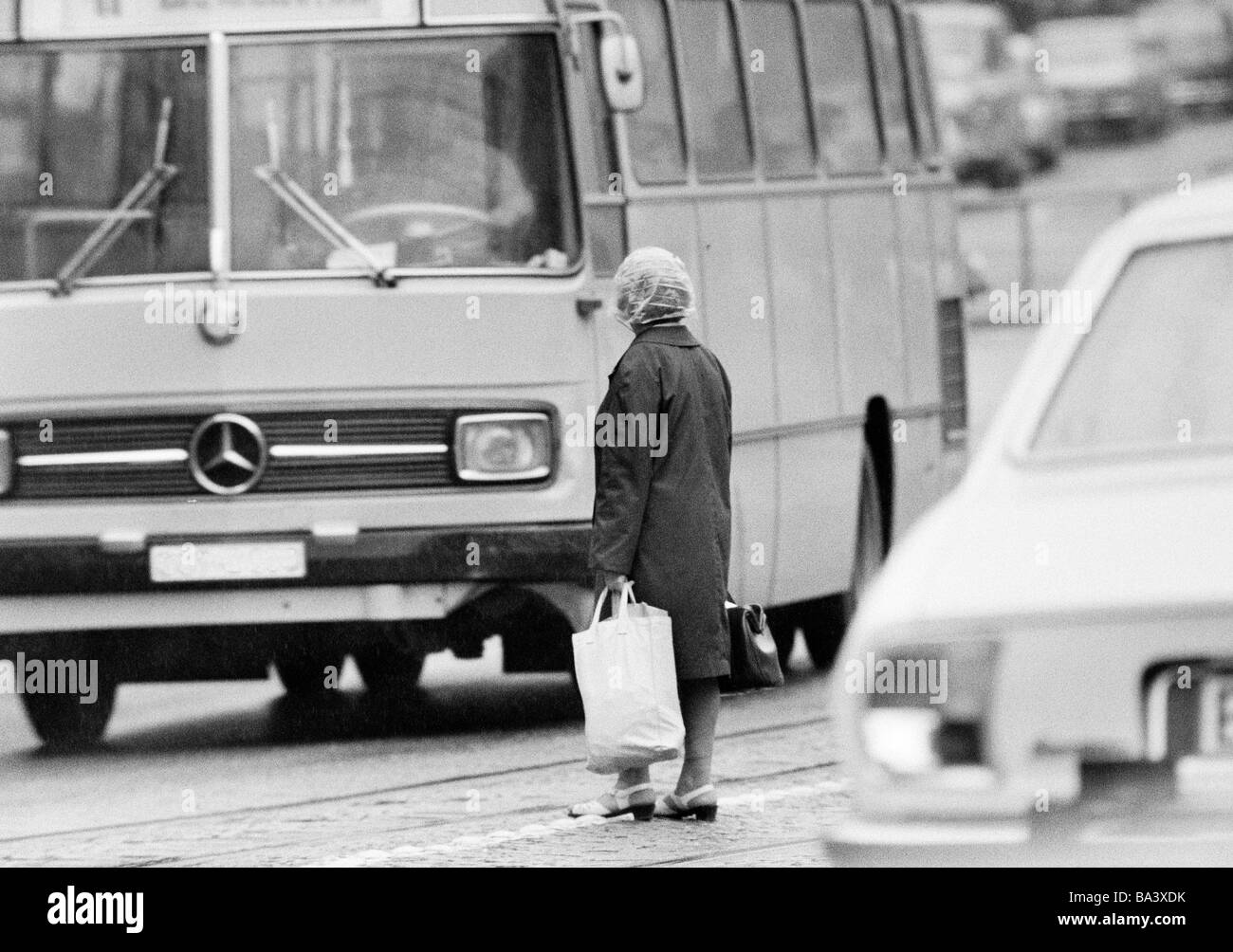 Seventies, black and white photo, people, older woman with shopping bags stands in the middle of a traffic road and tries to cross the road, heavy traffic, aged 60 to 80 years Stock Photo