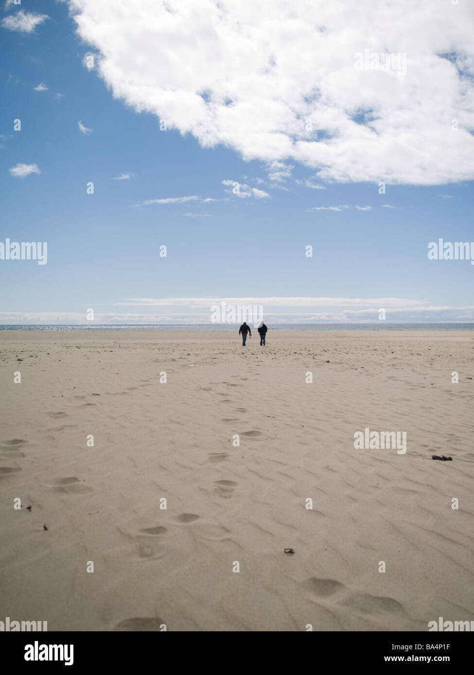 couple walking towards sea on long beach leaving footprints in the sand ...