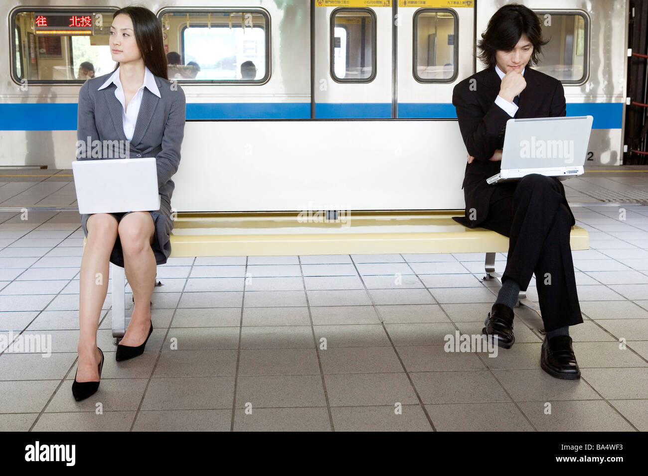 Businessman and businesswoman sitting on bench of train platform using laptops Stock Photo