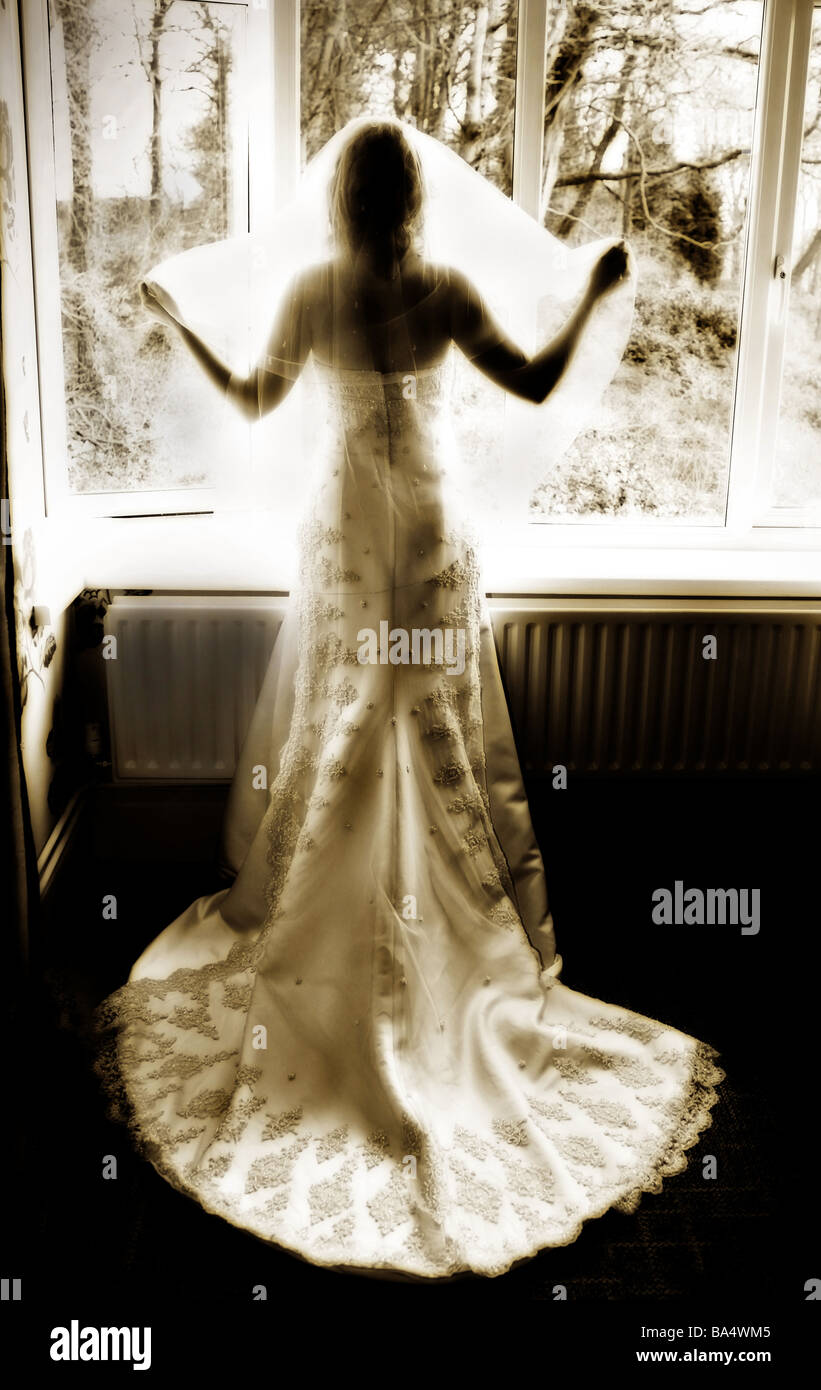 Silhouette of a bride in white wedding dress holding out wedding veil standing infront of window with woodland scene beyond Stock Photo